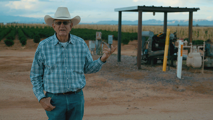 Man in a blue checked shirt with a cowboy hat stands in a green field with a well housing in the background.