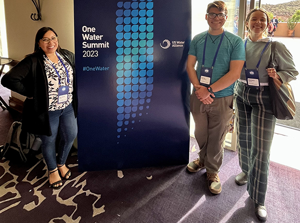 Three people stand in front of a banner reading "one water" in a conference center with purple carpeting and an outside patio in the background.