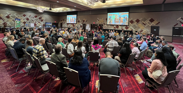 A crowd of people sit in a semi-round in a colorful conference room.