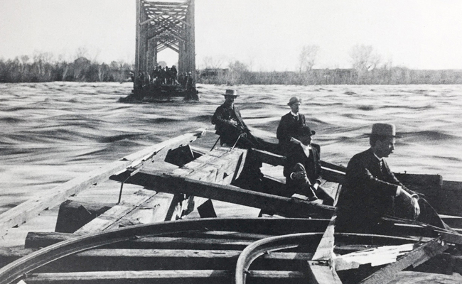 Men sit floating on a collapsed railroad bridge in a black and white image of a flooded river