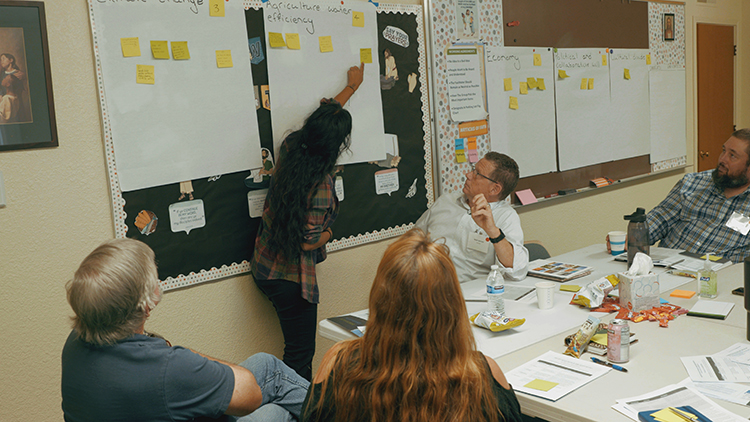 Four people sit facing a poster board filled with sticky notes as a woman with long dark hair points to one of the notes.