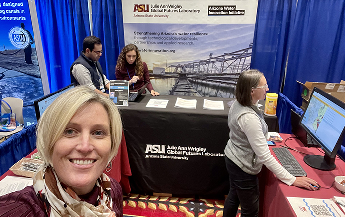 Four people stand in an exhibit booth at a conference, one taking a photo, another on a computer, and two looking at a document.