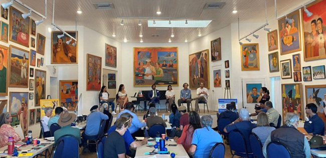 A room of people surrounded by colorful paintings listening to a panel discussion.