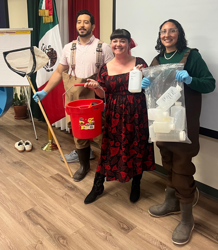 Three people stand with stream sampling equipment including a net and bucket