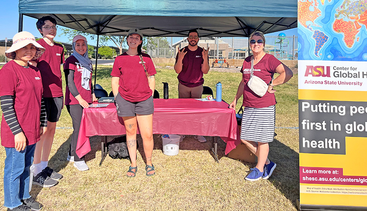 A group of people in ASU maroon t shirts stands at a table under a tent at an event.