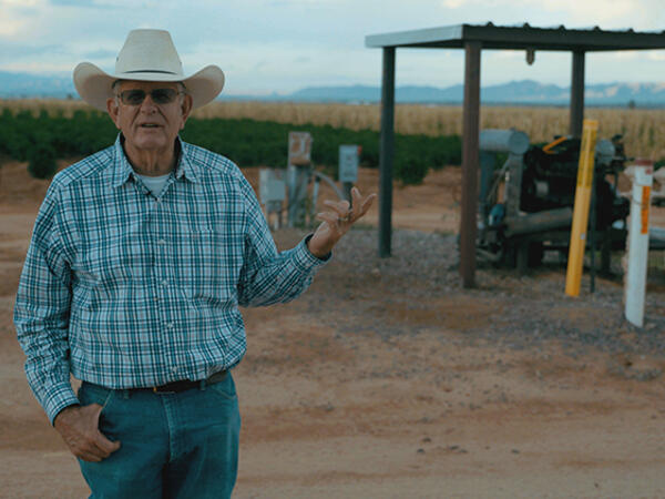 Man in a checked shirt stands in front of a well housing with field in background