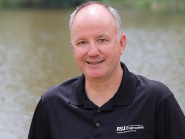 A man in a black polo shirt stands with a lake behind him.