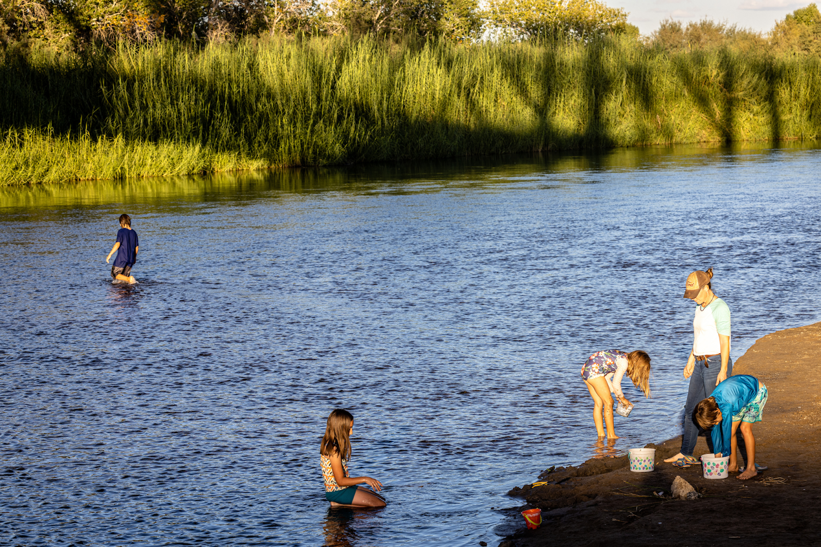  A blue river with green vegetation lining one side and a small beach on the other is the backdrop for four children wading the river and playing along the beach, along with their mother.