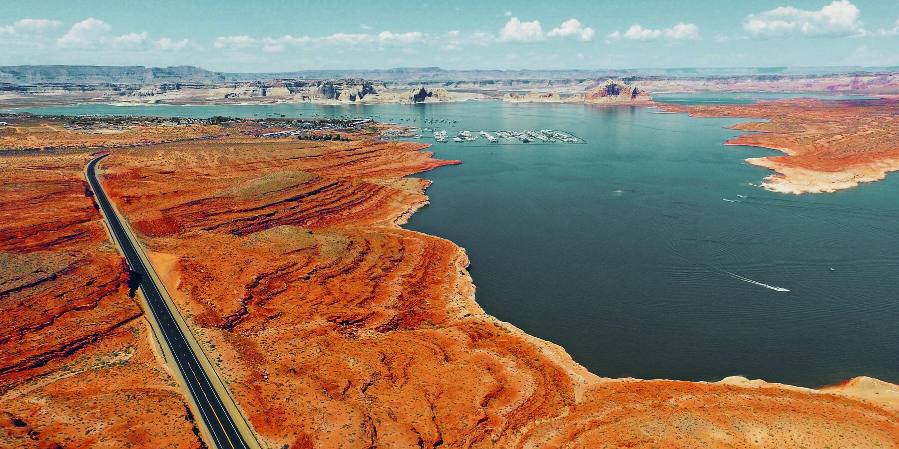 A large blue reservoir lake surrounded by red rock formations and a long road running alongside it glimmers in the sunlight. A marina shows in the far distance with blue skies and clouds.