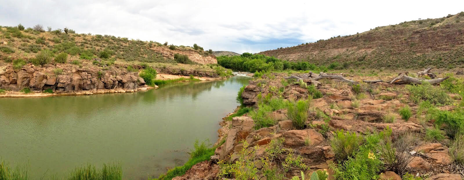 A river, the Verde River, flows through peach colored rock formations dotted with greenery and a cloudy sky overhead.