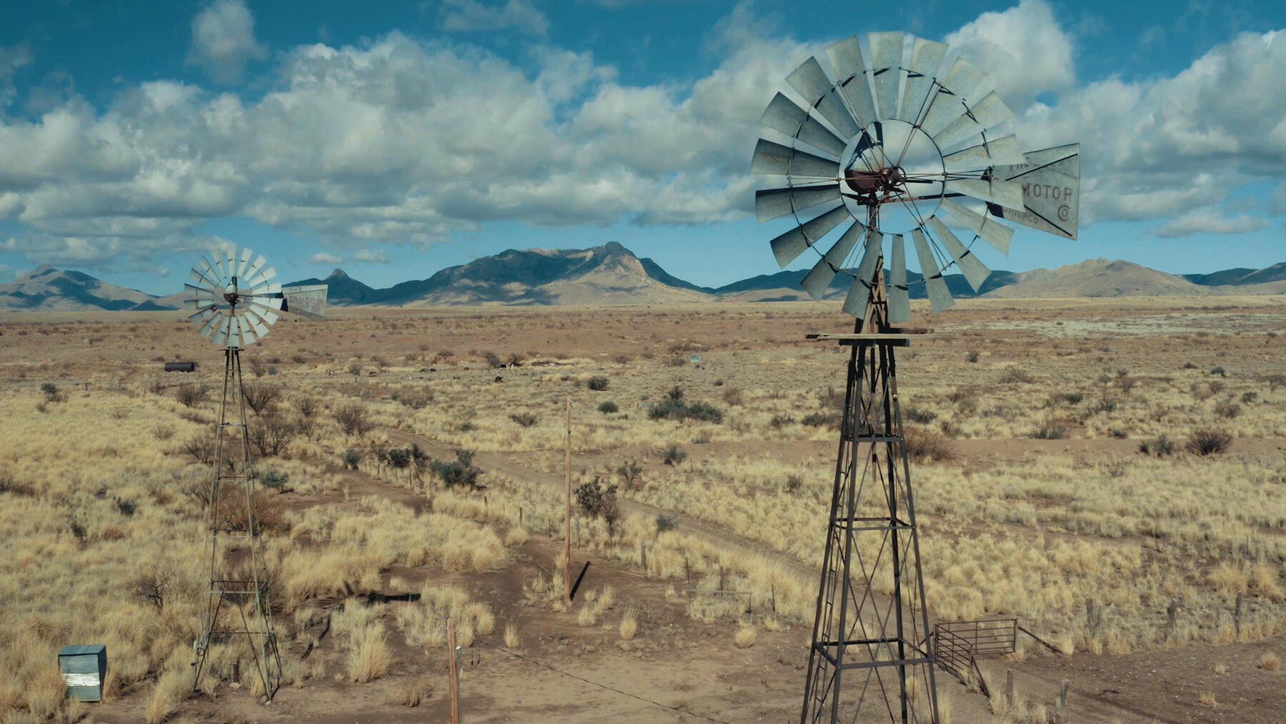 Two tall windmills stand in a landscape of desert grass and shrubs with mountains in the distance.