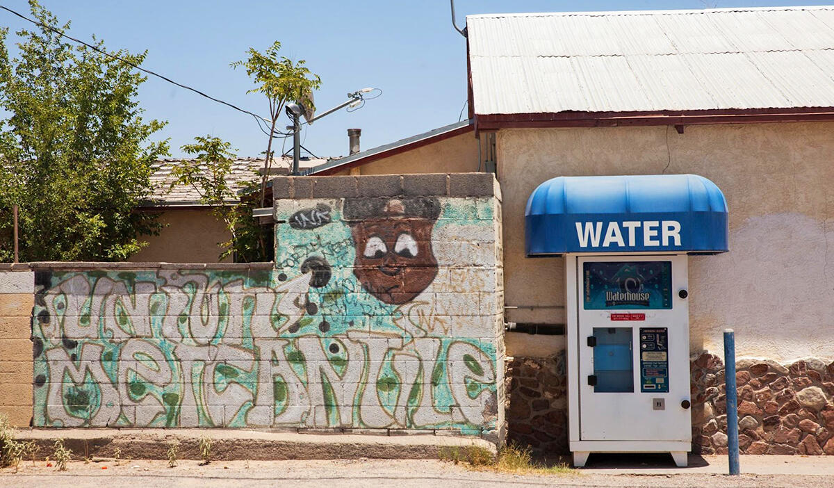 A water vending machine stands on a street with a bright mural and low desert building.