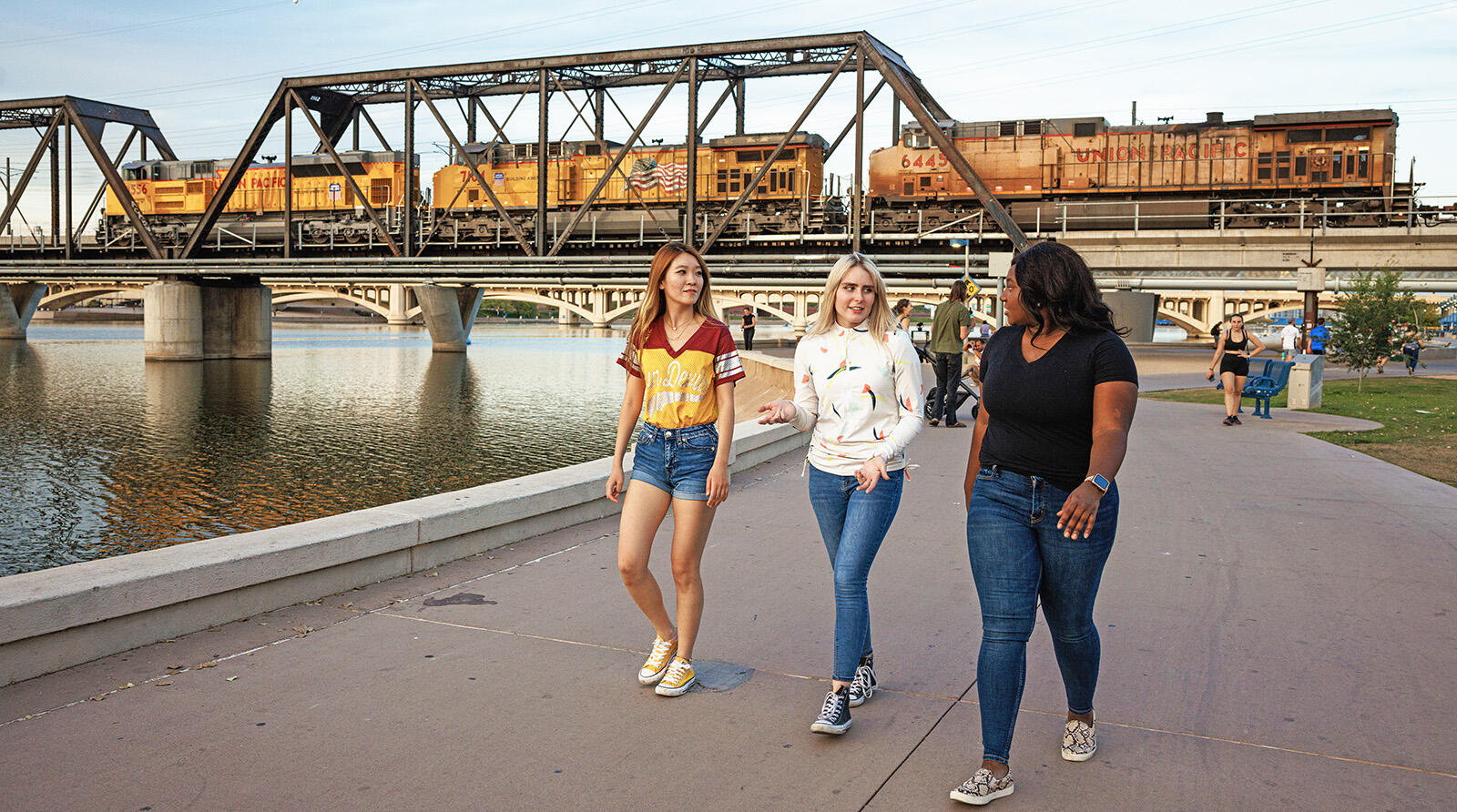 Three women walk along Tempe Town Lake with a train on the bridge in the background.