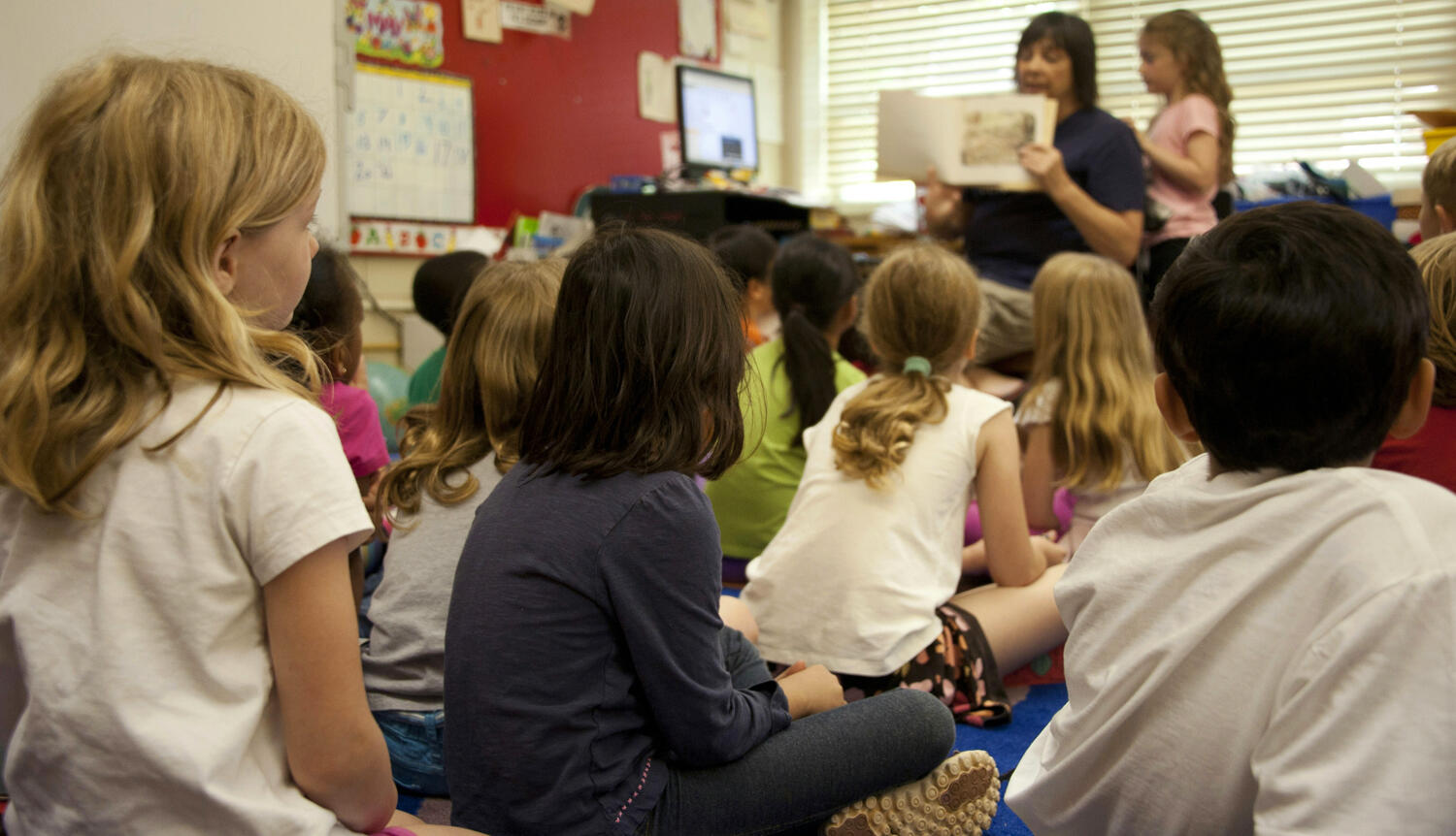 A group of children sit in front of an adult reading a book to them.