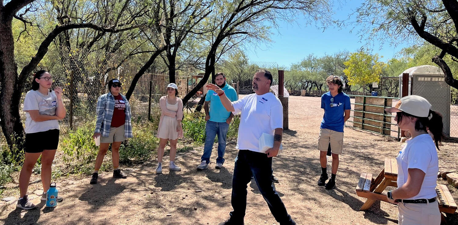 A group of people stand outside in the shade listening to a man in the center of a circle.