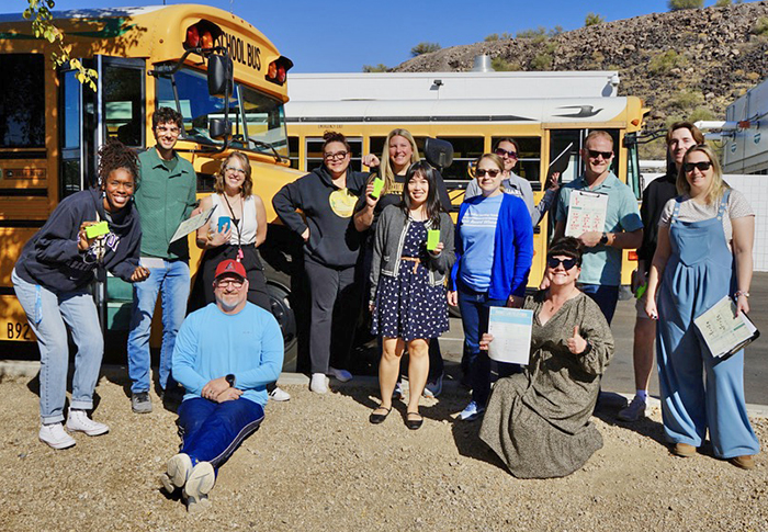 A group of people stand with devices outside a school bus